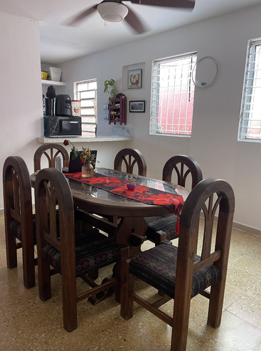 A cozy dining room features a wooden table with six chairs, a centerpiece with red flowers, kitchen appliances, wall art, and ample natural light from windows.
