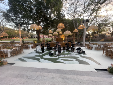 A band performs on a stylish outdoor stage surrounded by decorated tables and hanging lanterns, set amid trees and festive, elegant natural scenery at sunset.