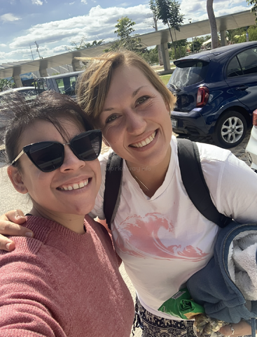 Two smiling women stand outside on a sunny day, taking a cheerful selfie. One wears sunglasses and a pink sweater, the other holds snacks and a jacket.