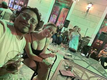 Ein fröhliches Paar genießt an einem Tisch im Freien ein paar Drinks und lächelt in die Kamera. Im lebhaften, stimmungsvollen Hintergrund des Lokals treten eine Live-Band und ein Sänger auf.