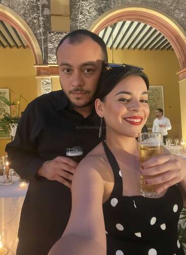 A smiling woman in a polka-dot dress and a man in black pose for a selfie, holding drinks at a warmly-lit, festive indoor event.