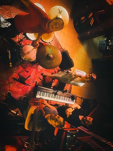 Four musicians stand together in a warmly lit room, surrounded by instruments including a drum set, keyboard, and bass guitar, smiling and looking up at the camera.