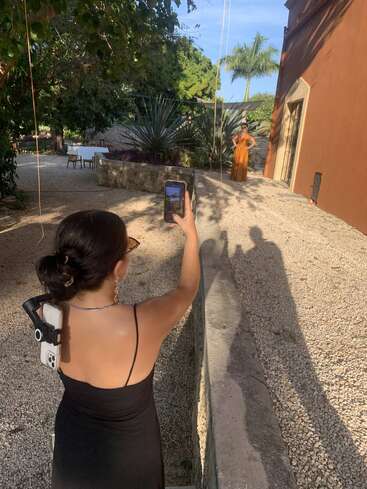 A woman in a black dress takes a photo of another woman in an orange dress outside, surrounded by trees, plants, and sunlight on a gravel path.