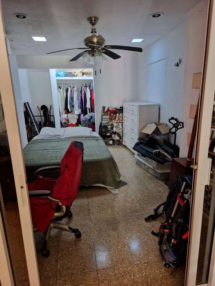A cozy bedroom with a ceiling fan, red desk chair, double bed, open closet, white dresser, various storage bins, and personal items organized throughout.