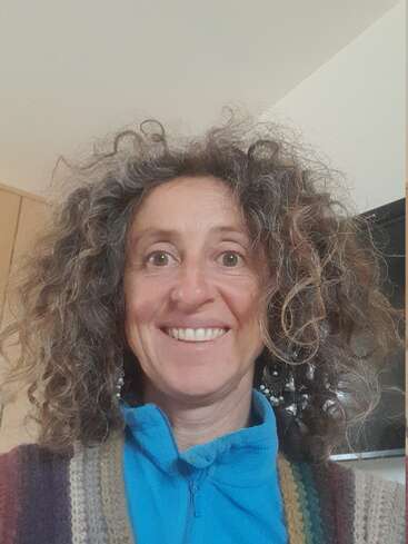 A woman with curly hair and a big smile is wearing a blue zip-up top and a colorful knitted sweater, standing indoors with a cheerful expression.