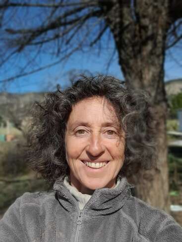 Das Bild zeigt eine Frau mit lockigem Haar und einer grauen Fleecejacke, die an einem sonnigen Tag im Freien vor einem Baum und einer Berglandschaft steht.