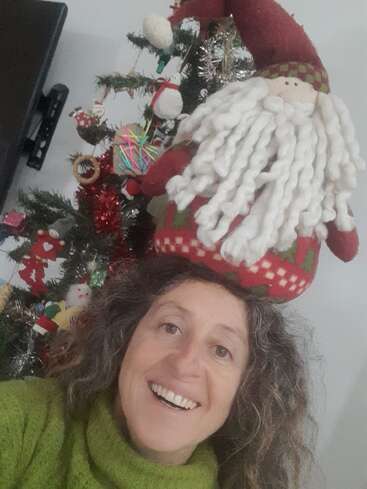 Una alegre mujer sonríe delante de un árbol de Navidad decorado, con un festivo jersey verde y un gorro de Papá Noel de felpa en la cabeza.