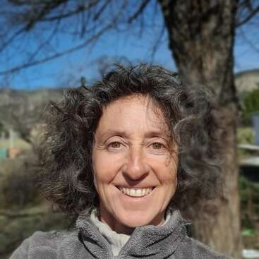 The image depicts a woman with curly hair and a gray fleece jacket, standing outdoors in front of a tree and a mountainous landscape on a sunny day.