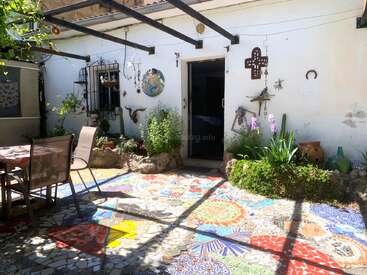 Ein lebhafter Innenhof mit Mosaikboden, Gartenmöbeln, üppigen Pflanzen und dekorativen Wandbildern, darunter ein Kreuz, ein Hufeisen und verschiedene Hängeornamente, im Sonnenlicht.