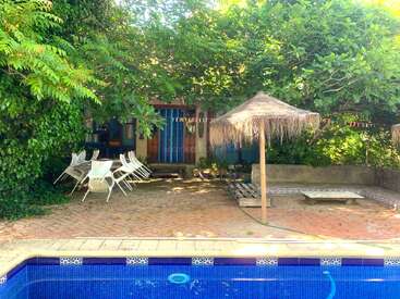 Das Bild zeigt eine gemütliche Terrasse mit Pool, die von üppig grünen Bäumen beschattet wird. Dort stehen weiße Stühle, ein Tisch und ein Sonnenschirm aus Stroh.