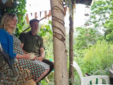 Eine Frau und ein Mann sitzen gemeinsam im Freien, umgeben von üppigem Grün. Sie wirken entspannt, halten Tassen in den Händen und sitzen unter einem rustikalen Unterstand, der mit hängenden Bändern und Windspielen geschmückt ist.