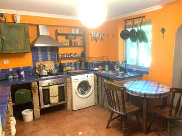 Diese gemütliche Küche besticht durch orangefarbene Wände, blau geflieste Arbeitsflächen, eine Waschmaschine, Holzmöbel, hängende Töpfe, offene Regale, diverse Küchenutensilien und eine karierte Tischdecke.