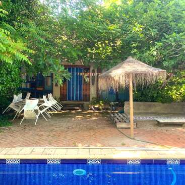 Esta imagen muestra un acogedor patio al aire libre con piscina, a la sombra de frondosos árboles. Hay sillas blancas, una mesa y una sombrilla de paja.