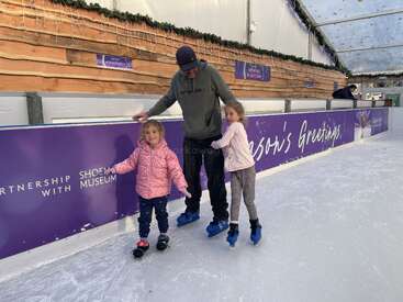 Un homme et deux jeunes filles patinent ensemble dans une patinoire couverte, souriant et se tenant à la barrière latérale décorée de signes festifs.