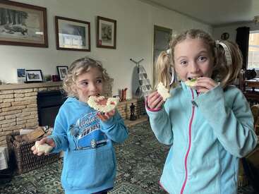 Deux jeunes filles aux cheveux bouclés se tiennent dans un salon confortable, mangeant joyeusement des sandwichs aux formes créatives, vêtues de sweats à capuche bleus et souriant avec des expressions espiègles et joyeuses.