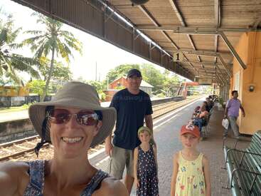 Une famille souriante pose sur le quai ensoleillé d'une gare, avec des palmiers en arrière-plan. D'autres passagers attendent sur des bancs et regardent vers l'appareil photo.