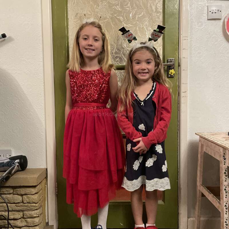 Deux jeunes filles se tiennent joyeusement à l'intérieur, vêtues de tenues de fête. L'une porte une robe rouge scintillante, l'autre une robe bleu marine à fleurs blanches et un bandeau avec un bonhomme de neige. Elles sourient radieusement.