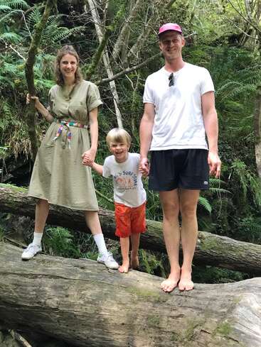 Eine dreiköpfige Familie steht lächelnd und händchenhaltend auf einem umgestürzten Baum in einem üppigen, grünen Wald. Das Kind ist barfuß und glücklich.