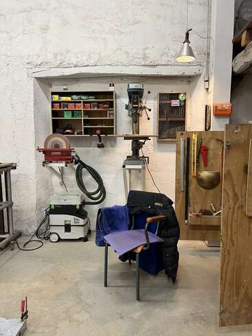 Eine aufgeräumte Werkstatt mit Elektrowerkzeugen, einer Wandbohrmaschine, geordneten Regalen mit Behältern, einem Stuhl mit Kleiderbügeln, einer Lampe und verschiedenen hängenden Werkzeugen.
