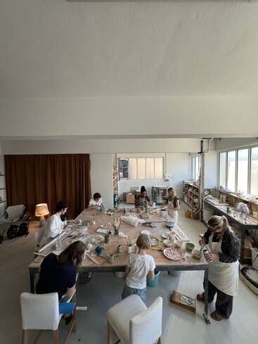 Sieben Personen, darunter ein Kind, arbeiten an Keramik um einen großen Tisch in einem hellen, gemütlichen Atelier, das mit Kunstzubehör, natürlichem Licht und Regalen gefüllt ist.