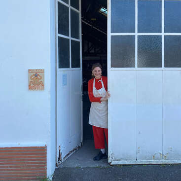 A smiling person in a red outfit and apron stands in the doorway of a white building labeled "Maison Meryveille," invitingly holding the large door open.