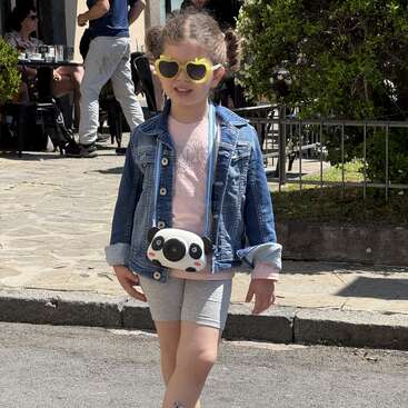 Ein junges Mädchen mit einer herzförmigen Sonnenbrille, einer Jeansjacke, einem rosa Shirt und grauen Shorts steht selbstbewusst draußen. Um den Hals hängt ihr eine süße Panda-Kamera.