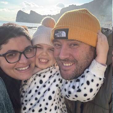 Une joyeuse famille de trois personnes sourit pour un selfie en gros plan en plein air, au bord de la mer. L'enfant porte une veste à pois et un bonnet. Un magnifique paysage.