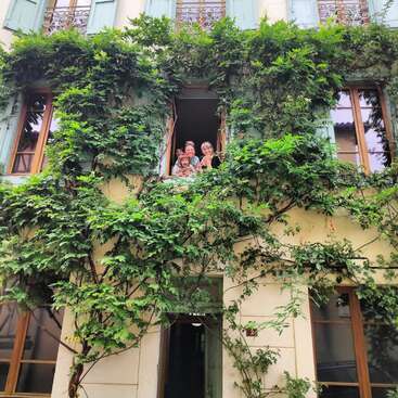 Das Bild zeigt ein charmantes Gebäude mit einem Balkon, der mit üppigem Grün bedeckt ist, und eine vierköpfige Familie, die aus dem Fenster schaut und in die Kamera winkt.