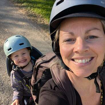A smiling woman and child wearing helmets take a cheerful selfie outdoors, likely during a bike ride on a sunny day, enjoying time together and nature.
