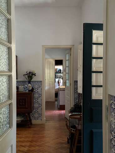 Sunlight streams into a cozy hallway with parquet floors, blue patterned tiles, and open doors leading to a kitchen where a woman works at the counter.