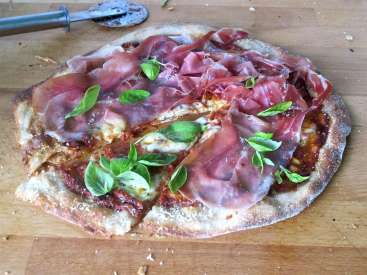 La imagen muestra una pizza parcialmente consumida con hojas de albahaca y jamón serrano sobre una tabla de cortar de madera, acompañada de un cortador de pizza.
