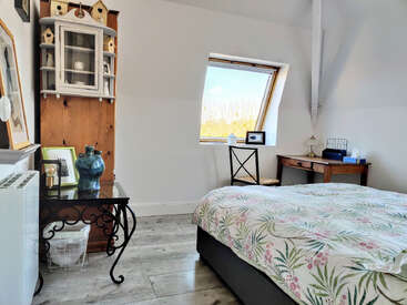 Un acogedor dormitorio con ropa de cama a flores, suelos de madera, un escritorio junto a la ventana, una mesita auxiliar negra, un jarrón azul, un armario con forma de pajarera y luz natural.