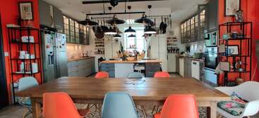Una cocina amplia y moderna con paredes naranjas, una mesa de comedor de madera, sillas de colores, lámparas colgantes, estantes metálicos, vitrinas y utensilios de cocina bien ordenados en la isla central.