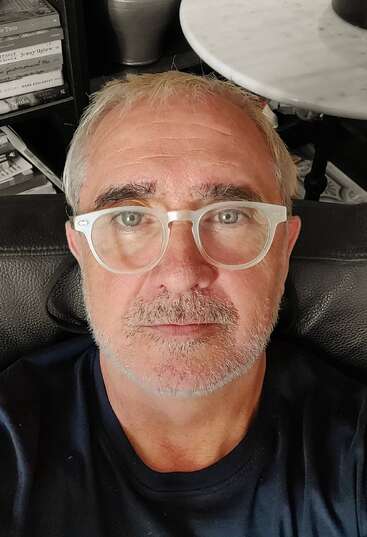 Un hombre de mediana edad, con el pelo y la barba canosos y gafas transparentes, mira directamente a la cámara. Detrás de él hay estanterías y una mesa redonda blanca.