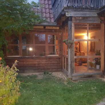 Cette image montre une maison en bois chaleureuse et rustique, dotée de grandes fenêtres, d'un intérieur baigné d'une lumière tamisée, d'un cadre verdoyant à l'extérieur et d'une atmosphère paisible en fin de journée qui invite à la chaleur et à la détente.