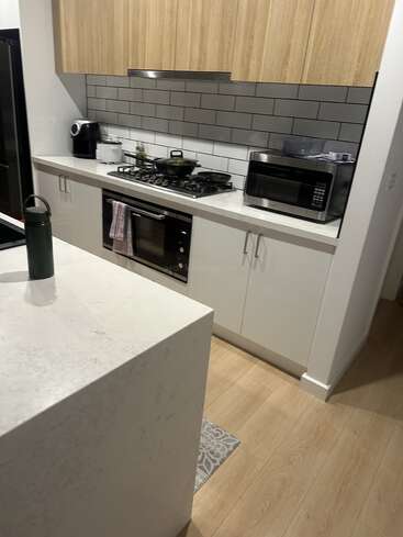 Cozinha moderna com armários de madeira, bancadas brancas, fogão a gás, forno, micro-ondas, chaleira e fritadeira. Backsplash de azulejos cinza, garrafa de água, ilha e piso de madeira. Limpa e organizada.