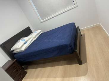 Esta imagem mostra um quarto minimalista com uma cama de madeira escura, roupa de cama azul, dois travesseiros, uma pequena mesa de cabeceira, piso de madeira clara e uma janela.