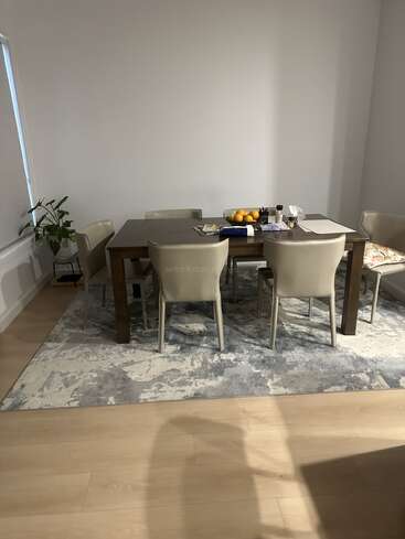 Uma sala de jantar moderna apresenta uma mesa de madeira, seis cadeiras bege, um tapete estampado, um vaso de plantas, uma fruteira, revistas e uma decoração minimalista.