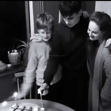 Three people, two boys and a woman, are standing together smiling, about to cut a cake with lit candles, suggesting a celebratory, heartfelt moment in black and white.