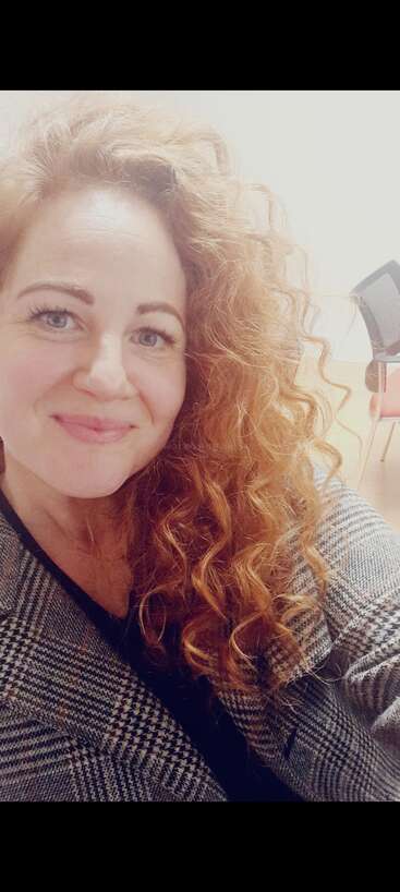 A woman with curly red hair smiles at the camera, wearing a checked coat. The background is light with a chair visible, giving a warm atmosphere.