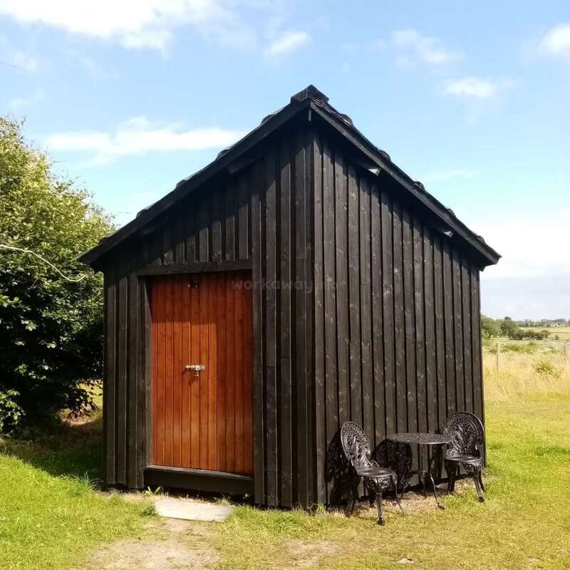 A small, dark wooden shed with a brown door stands on green grass. Two ornate black metal chairs and a table sit outside under a blue sky.
