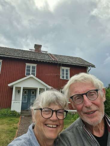 Um casal de idosos com cabelos grisalhos e óculos sorri em frente a uma grande e tradicional casa de madeira vermelha sob um céu nublado, exalando felicidade e cordialidade.