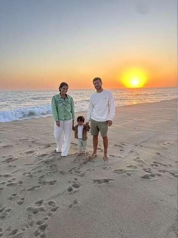 Uma família de três pessoas está descalça na areia da praia ao pôr do sol. O sol se põe sobre o oceano, criando uma atmosfera calorosa, pacífica e alegre.