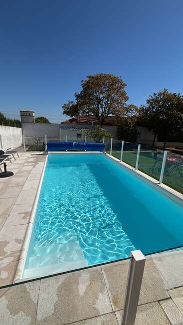 Uma piscina retangular azul cintilante com água limpa, cercada por uma cerca de vidro, espreguiçadeiras e um pátio iluminado pelo sol, sob um céu azul sem nuvens.