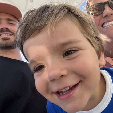 Uma alegre selfie de família mostra um menino em primeiro plano sorrindo amplamente, com um homem e uma mulher atrás dele, ambos sorrindo alegremente para a câmera.