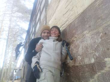 Um casal sorridente posa em frente a uma parede de madeira, bem agasalhado, com um deles segurando uma ferramenta. Outra pessoa aparece trabalhando em uma escada nas proximidades.