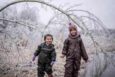 Duas crianças com roupas quentes e à prova d'água estão ao ar livre, cercadas por galhos de árvores curvados e cobertos de gelo. A atmosfera é invernal, com as duas crianças parecendo satisfeitas e aventureiras.