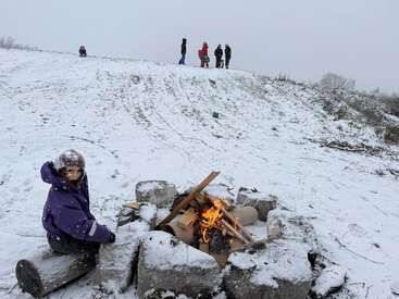 Uma criança com uma jaqueta roxa se aquece perto de uma pequena fogueira em uma área externa com neve. As crianças estão andando de trenó e brincando em uma colina com neve ao fundo.