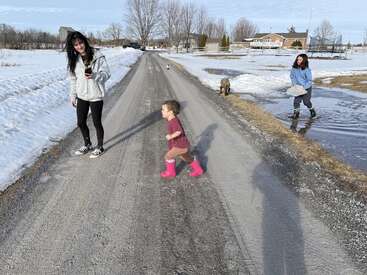 Uma criança com botas cor-de-rosa corre em uma estrada rural coberta de neve, enquanto duas mulheres e um cachorro aproveitam o degelo da primavera em um ambiente rural tranquilo.