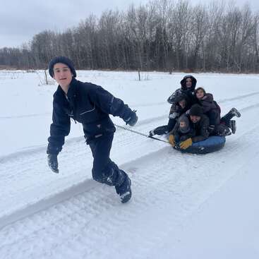A person dressed in winter clothes pulls a snow tube filled with five smiling children across a snowy field, with bare trees in the background. Joyful winter fun.